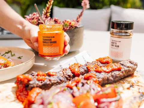 A jar of Turkish Chilli Sauce being held by a hand, with the sauce applied to a dish in the foreground.
