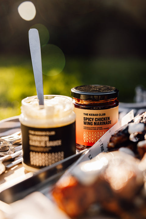 Two jars of marinade with a spoon on a grill, set against a blurred outdoor background.