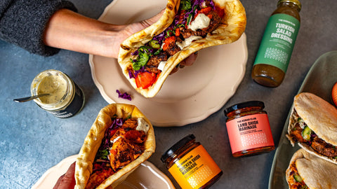 Person holding a pita with salad, surrounded by various condiments on a textured surface.