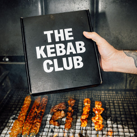 Person holding a 'The Kebab Club' box with skewered kebabs on a grill.
