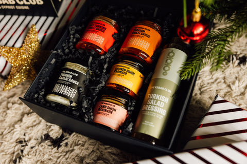 A gift box containing six various marinades and sauces, with decorative elements like a Christmas tree and ornaments visible in the background.