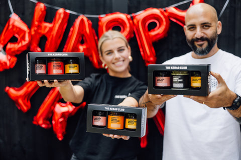 Two people holding The Herban Club product boxes with red 'HAPPY' balloons in the background.